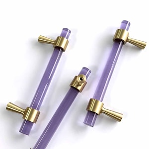 Può includere: Tre maniglie per armadietti con barre cilindriche viola traslucide e finiture in metallo oro spazzolato. Ogni maniglia presenta due supporti conici, con una maniglia che mostra un punto di fissaggio centrale sul collare metallico.