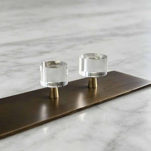 May include: Two cylindrical cabinet knobs featuring clear, transparent acrylic tops mounted on small, tapered metallic gold bases. The knobs are attached to a rectangular, brushed bronze-toned metal plate, showcasing a clean and functional design.