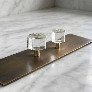 May include: Two cabinet knobs featuring clear, faceted, hexagonal glass tops mounted on conical, brushed brass-toned metal bases. The knobs are attached to a rectangular, brushed brass-toned metal plate, showcasing a clean and functional design.