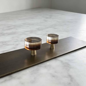 May include: Two circular cabinet knobs featuring a translucent amber and brown gradient resin top mounted on a brushed brass conical base. The knobs are attached to a rectangular, dark bronze-toned metal plate with a smooth, matte finish.