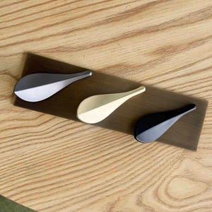 May include: Three teardrop-shaped cabinet knobs mounted on a rectangular display board. The knobs feature a smooth, solid finish in three distinct colors: a metallic silver-grey, a brushed gold, and a matte black, all arranged in a diagonal row.