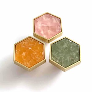 May include: Three hexagonal cabinet knobs with gold-toned metal frames. Each knob features a flat, marbled inlay surface in distinct colors: one soft pink, one vibrant orange, and one muted sage green, all displaying a textured, cloudy pattern.