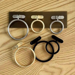 May include: A collection of five circular metal cabinet pulls in various finishes including brushed silver, gold, and matte black. Three pulls are mounted on a rectangular bronze-colored plate, while two are loose, featuring a ring and a rectangular base.