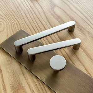 May include: Two brushed silver metal drawer pulls with a long, rectangular bar design and rounded ends, accompanied by a single matching circular knob. These hardware pieces are mounted on a flat, rectangular, brushed brass-toned metal plate.