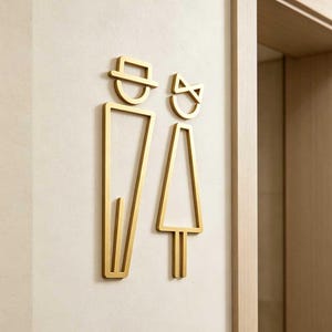 Könnte beinhalten: Goldfarbene Toilettenschilder, die einen Mann und eine Frau darstellen. Das Schild des Mannes hat einen Hut, das der Frau eine Schleife. Die Schilder sind an einer hellen Wand in der Nähe eines Holzrahmen angebracht.