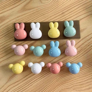 Puede incluir: Una colección de once pomos para armarios pequeños, brillantes y de aspecto cerámico en varios colores pastel, incluyendo rosa, blanco, amarillo, azul y verde menta. Los pomos tienen forma de cabeza de conejo con orejas largas o redondas.