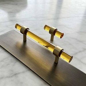 May include: A long cabinet pull and a smaller matching cabinet knob, both featuring a cylindrical, translucent amber-coloured acrylic bar held by brushed brass metal mounts, mounted on a rectangular brushed brass metal plate.