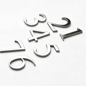 May include: A collection of seven individual silver-colored metal house numbers, specifically the digits 1, 2, 3, 4, 5, 6, and 7. Each numeral features a clean, sans-serif typeface with a smooth, reflective metallic finish and a three-dimensional profile.