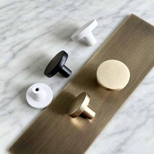 May include: A collection of round cabinet knobs in white, matte black, and brushed brass finishes. The knobs feature a simple cylindrical base and a flat circular top. One white knob is shown upside down, revealing the central screw hole.