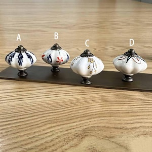 Pode incluir: Quatro puxadores de armário em cerâmica branca com formato canelado semelhante a uma abóbora e bases de metal escuro. Cada puxador apresenta um design único pintado à mão: A tem folhas azuis, B motivos florais vermelhos e azuis, C flores douradas e D padrões verdes e vermelhos.