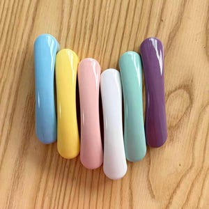 May include: A set of six smooth, elongated cabinet handles in a variety of glossy colors including light blue, yellow, soft pink, white, mint green, and deep purple. Each handle features a curved, rounded shape with a polished finish.