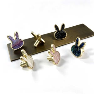 May include: A collection of five rabbit-shaped cabinet knobs with gold-toned metal edges. Each knob features a unique inlaid centre, including purple, white, black with glitter, light pink, and blue-green marbled patterns, with a cylindrical base.