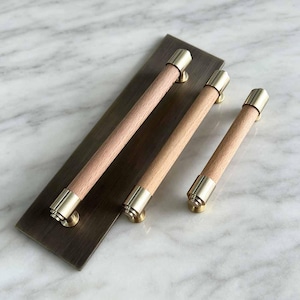 May include: Three cylindrical cabinet handles featuring light-toned wooden grips and polished brass end caps. Two of the handles are mounted onto a rectangular, brushed bronze-toned metal backplate, while the third handle is separate.