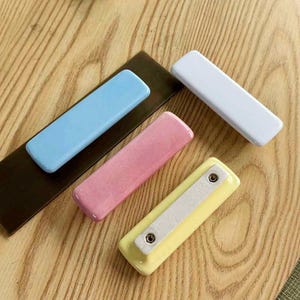 May include: Four rectangular cabinet pulls in pastel colors including light blue, white, pink, and yellow. The light blue pull is attached to a dark brown rectangular base, while the yellow pull shows the underside with two metal screw holes.