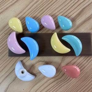 May include: A collection of ceramic cabinet knobs in pastel colours, including yellow, blue, pink, and light blue. The knobs are shaped like teardrops and crescent moons, featuring a smooth, glossy finish and a metal mounting point on the back.