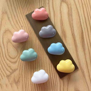 May include: A collection of seven cloud-shaped knobs in various pastel colours including pink, light blue, mint green, yellow, white, and grey. These smooth, glossy cabinet pulls are arranged in a cluster, with some attached to a rectangular strip.