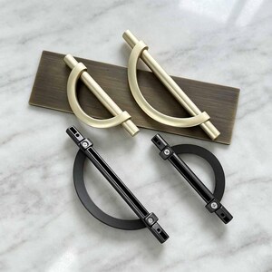 May include: Two matte gold semi-circular cabinet pulls resting on a rectangular bronze plate, alongside two black semi-circular cabinet pulls with visible mounting screws. The hardware features a curved arch design connected to a straight horizontal bar.