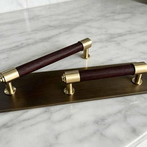 May include: Two cabinet pulls featuring dark brown wooden cylindrical handles with brushed brass metal end caps and mounting posts, attached to a rectangular brushed brass backplate with a metallic finish and visible horizontal grain.