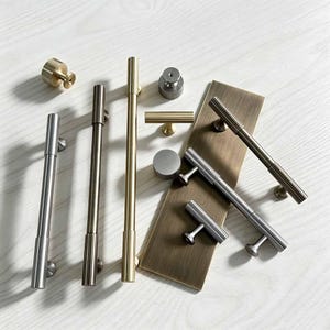 May include: A collection of metal cabinet hardware including long cylindrical pulls, T-bar handles, and round knobs in brushed gold, silver, and bronze finishes. Some hardware pieces are arranged on a rectangular brass-toned metal plate.
