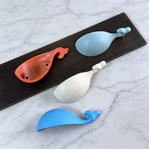 Peut inclure: Un ensemble de quatre poignées de tiroir en forme de baleine, déclinées en orange, bleu clair, crème et bleu. Chaque poignée présente un corps lisse et incurvé avec une queue, ainsi que deux trous de vis circulaires pour la fixation.