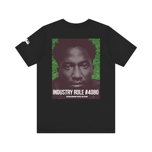 May include: Black t-shirt with a portrait of a person set against a green backdrop. The text "INDUSTRY RULE #4080" and "RECORD COMPANY PEOPLE ARE SHADY" are printed below the portrait. The shirt has short sleeves.
