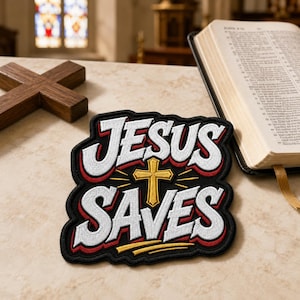 May include: A black embroidered patch featuring the white text JESUS SAVES with a gold cross in the center. The lettering has a red outline and a black border. The patch is shaped to follow the contour of the text and the central cross design.