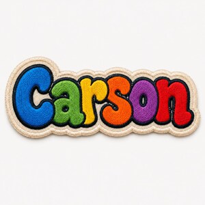 May include: An embroidered patch featuring the name Carson in a bubbly, rounded font. Each letter is a different color: blue, green, yellow, orange, purple, and red. The entire word is outlined in black and set against a cream-colored fabric border.