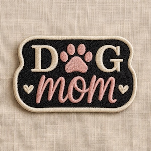 May include: Embroidered patch with the words "DOG MOM" in cream and pink script, featuring a pink paw print and two small hearts. The patch has a black background and a cream-colored border.