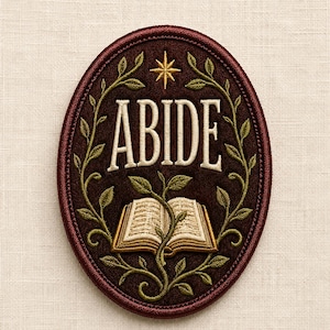 May include: An oval-shaped embroidered patch featuring a dark brown background with a cream-colored open book at the center. The word ABIDE is stitched in cream above the book, surrounded by green vines and a gold star at the top border.