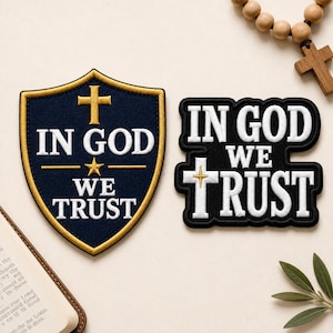 May include: Two embroidered patches featuring the phrase IN GOD WE TRUST. One patch is a navy blue shield with gold trim, a gold cross, and a gold star. The second patch is black with white lettering and a white cross featuring a small gold star detail.