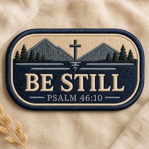 May include: An oval embroidered patch featuring a dark blue border and a landscape scene with grey mountains, dark green pine trees, and a central cross. The text "BE STILL" and "PSALM 46:10" are stitched in cream-colored thread on a dark blue background.