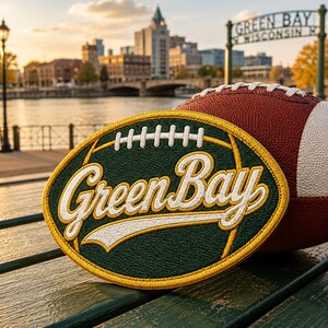Puede incluir: Un bal&oacute;n de f&uacute;tbol americano marr&oacute;n y blanco descansa junto a un parche bordado en verde y dorado. El parche dice "Green Bay" en letras blancas. El fondo muestra un paisaje urbano y un letrero que dice "Green Bay Wisconsin".