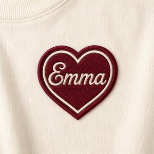 May include: Cream-colored sweatshirt with a burgundy heart-shaped patch. The patch has a white border and the name "Emma" embroidered in a cursive font. The sweatshirt has a ribbed neckline.