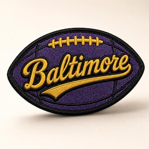 May include: Embroidered patch in the shape of a football, featuring the word "Baltimore" in gold script. The patch has a purple background with black stitching around the edges and details.