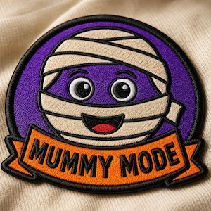 May include: A circular embroidered patch featuring a cartoon mummy face with large eyes and a smiling mouth wrapped in beige bandages against a purple background. Below the face is an orange banner with the black text MUMMY MODE.