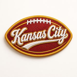May include: Embroidered patch in the shape of a football, with a maroon background and gold trim. The words "Kansas City" are in white script across the center. White stitching details the laces.