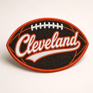 May include: Embroidered patch in the shape of a football, featuring the word "Cleveland" in white script with an orange outline. The patch has a black background with orange and white details.