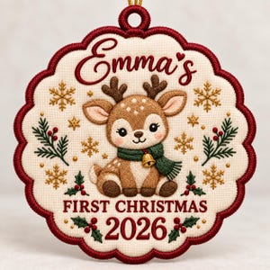 May include: A scalloped, cream-colored embroidered ornament featuring a brown fawn wearing a green scarf with a small bell. The text reads Emma's FIRST CHRISTMAS 2026. The design includes gold snowflakes, green pine sprigs, and red holly berries.