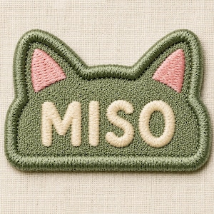 May include: Embroidered patch in the shape of a cat's head, with the word "MISO" in cream-colored thread. The cat head is olive green with pink inner ears. The patch has a textured appearance and a stitched border.