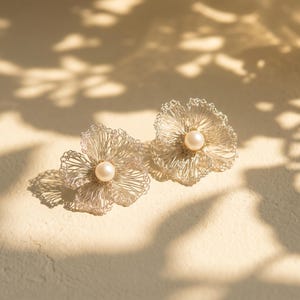 Pode incluir: Um par de brincos delicados em forma de flor. Cada brinco apresenta um design semelhante a arame em prata clara, com uma pérola no centro. Os brincos estão sobre um fundo macio e neutro com sombras.