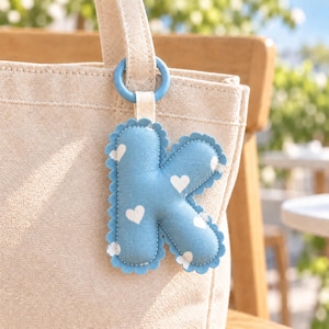 Può includere: Un ciondolo per borsa a forma di lettera K imbottita in tessuto azzurro con un motivo a cuori bianchi. La lettera presenta un bordo smerlato con cuciture visibili ed è fissata a un anello circolare azzurro tramite un piccolo passante in tessuto.