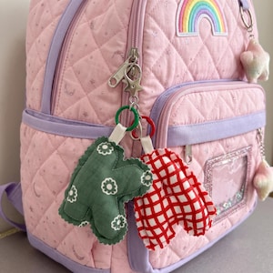 May include: A light pink quilted backpack with a rainbow patch and star-shaped zipper pulls. Two plush letter charms hang from the zipper: a green letter K with a white floral pattern and a red and white checkered letter A, both attached by rings.
