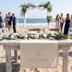 Puede incluir: Mesa de recepción de boda en la playa, con un camino de mesa de lino crema con los nombres "Declan & Sloane" y la fecha 20.07.2026. La mesa está adornada con rosas blancas, vegetación, conchas marinas y velas. Se ve un arco de madera.