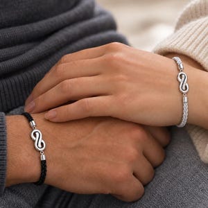 Könnte beinhalten: Zwei geflochtene Kordelarmbänder mit einem silberfarbenen Unendlichkeitssymbol als Mittelstück. Ein Armband besteht aus einer schwarzen Kordel, das andere aus einer weißen Kordel. Beide Unendlichkeitsanhänger sind mit kleinen, klaren Steinen besetzt.