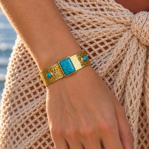 Könnte beinhalten: Ein goldfarbenes Armband mit einem zentralen rechteckigen Türkisstein und zwei kleineren runden Türkissteinen an den Seiten. Die Metalloberfläche des Armbands ist über die gesamte Länge mit einem komplizierten, geprägten Muster verziert.