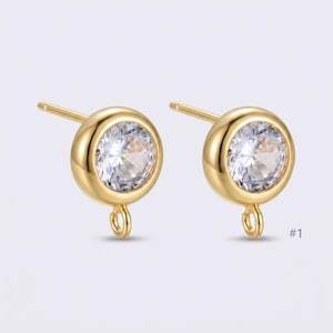 Puede incluir: Dos pendientes dorados con una piedra clara, redonda y facetada en el centro. Cada pendiente tiene un poste para insertar y un pequeño lazo en la parte inferior. El fondo es de color claro.