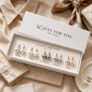 May include: A white rectangular gift box labeled JUST FOR YOU BEST WISHES containing five pairs of pearl earrings arranged by day of the week from Monday to Friday. The earrings feature gold-tone hoops with white, grey, and cream-colored pearls.