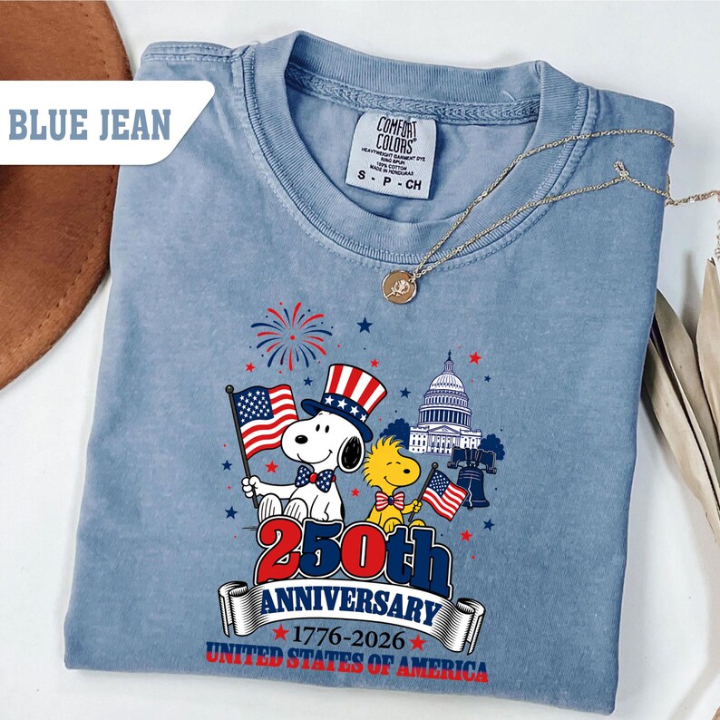 May include: Blue jean colored t-shirt featuring Snoopy and Woodstock with American flags, a patriotic hat, and the Capitol building. The shirt commemorates the 250th Anniversary of the United States of America, 1776-2026.