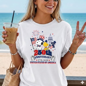 May include: White t-shirt featuring Snoopy and Woodstock with American flags, celebrating the 250th Anniversary of the United States of America. The design includes the Capitol building, fireworks, and the text "250th Anniversary 1776-2026 United States of America."