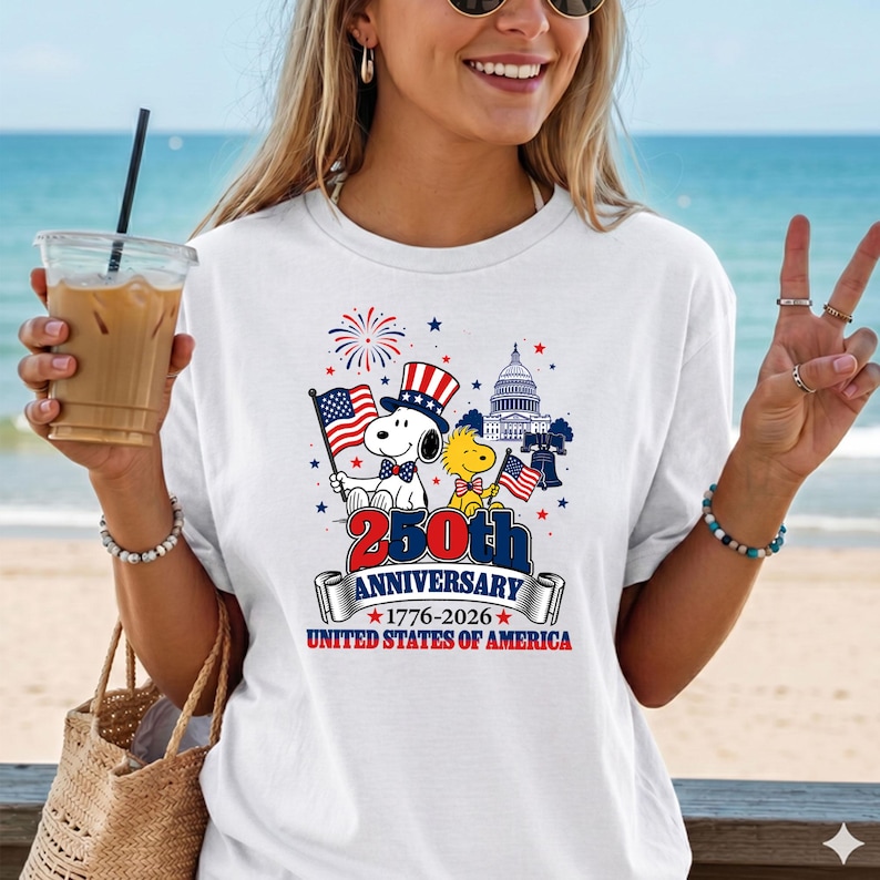 May include: White t-shirt featuring Snoopy and Woodstock with American flags, celebrating the 250th Anniversary of the United States of America. The design includes the Capitol building, fireworks, and the text "250th Anniversary 1776-2026 United States of America."